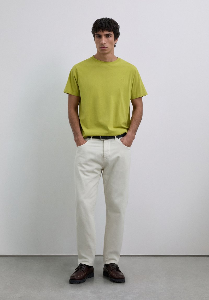 Homme debout avec les mains dans les poches, portant un t-shirt vert lime, un pantalon blanc cassé, une ceinture noire et des chaussures brun foncé devant un mur uni.