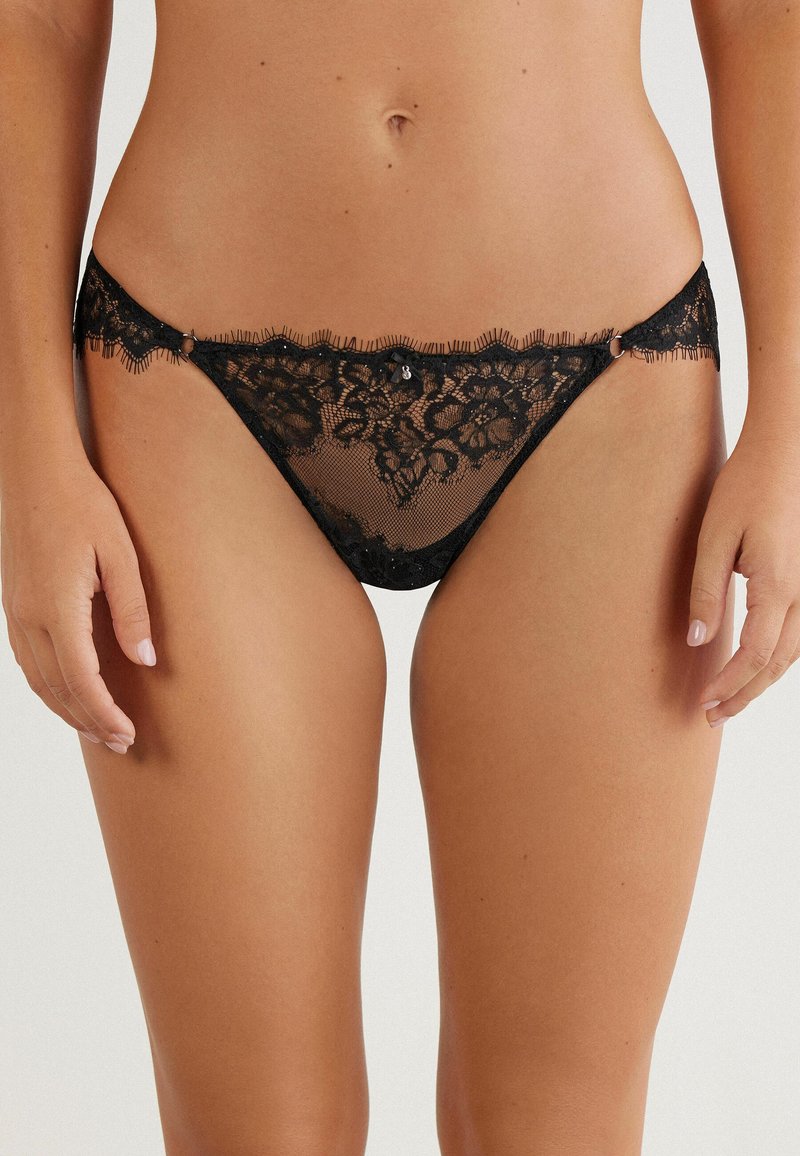 Zwarte kant tang met bloemenpatroon, doorschijnende mesh voorkant, golvende randen en decoratieve hardware-accenten aan de zijkanten. Zachte, delicate textuur.