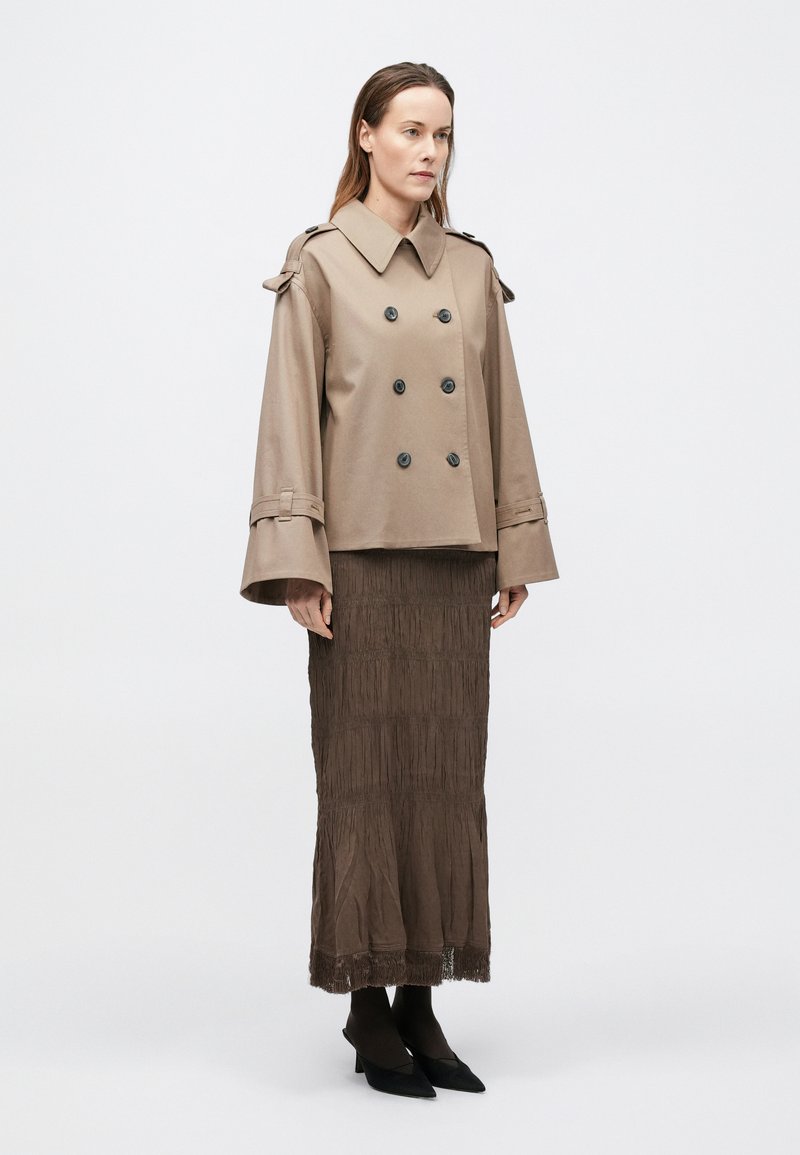 Manteau beige croisé avec de gros boutons, manches larges et épaulettes, associé à une jupe longue plissée marron avec des détails frangés.