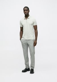 Gaiši pelēks polo krekls ar īsu rāvējslēdzi, valkāts kopā ar gaiši pelēkām biksēm un melniem apaviem. Vienkāršs dizains, gluda tekstūra, minimāls zīmola noformējums.