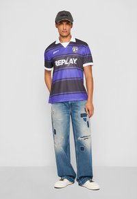 Black and purple striped jersey with "REPLAY" logo, white collar, paired with distressed light blue wide-leg jeans and white sneakers.