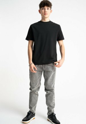 Młody mężczyzna stoi, ubrany w czarną koszulkę, szare dżinsy, czarne trampki oraz srebrną bransoletkę na lewej ręce, na białym tle.