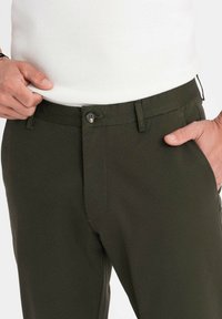 Mörkgröna byxor i texturerat tyg har en knappstängning, bältesöljar och ett platt framstycke, delvis synliga vid midjan.