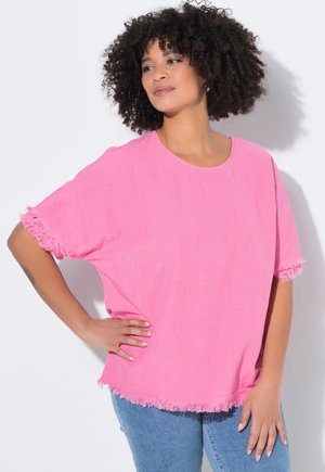 Kvinde med krøllet hår iført en knaldrød pink kortærmet top med frynser og blå jeans, poserer med den ene hånd på hoften og kigger til siden.