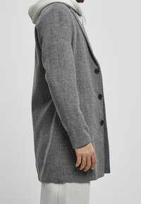 Grå fiskebensmønstret frakke med en struktureret design, fem knapper og et tekstureret stof. Hættetrøje synlig under.