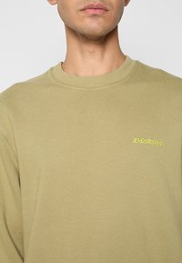 Homme portant un sweat-shirt à col rond vert olive clair avec un petit logo jaune "Dickies" sur le côté gauche de la poitrine.