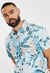 Camicia a maniche corte di colore azzurro chiaro con motivo floreale tropicale in rosa e verde. Presenta una chiusura frontale con bottoni e una vestibilità rilassata.