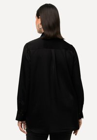 Maglietta nera a maniche lunghe realizzata in tessuto morbido, caratterizzata da un colletto, polsi abbottonati e una vestibilità leggermente ampia. Il retro presenta un dettaglio con cucitura centrale.