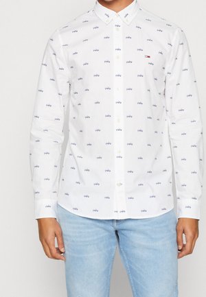 Biała koszula na guziki z małymi niebieskimi nadrukami rowerów i małym logo Tommy Hilfiger na piersi, noszona z jasnoniebieskimi dżinsami.