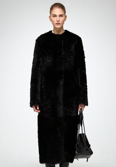 Cappotto in pelliccia sintetica nera con vestibilità ampia, maniche lunghe e design fino al ginocchio. La modella tiene in mano una borsa a tracolla nera e struttura rigida.