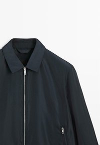 Sort lynlåstrøje med krave og lynlåslomme i siden, vist mod en hvid baggrund.