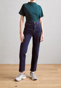 Zelený krátký rukáv, vysoký pas tmavě modré džíny, šedé sportovní boty. Hladká tkanina, rovný střih, jemné detaily prošití.