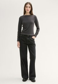 Maglia maniche lunghe di colore grigio scuro realizzata in un tessuto morbido, abbinata a jeans neri a gamba larga a vita alta. Il modello indossa scarpe nere, con le mani in tasca.