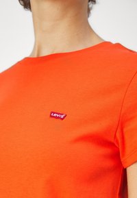 Gros plan d'une personne portant un t-shirt Levi's orange vif avec un petit logo Levi's rouge et blanc sur la poitrine.