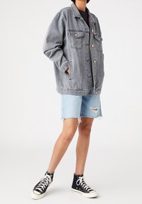 Grå denim oversized jacka med synlig sömnad och tryckknappar, kombinerad med ljusblå slitna denimshorts och svarta högskurna sneakers.