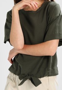 Olive green short-sleeve top with a rounded neckline, knotted detail at the waist, and soft fabric texture. Simple, relaxed fit.