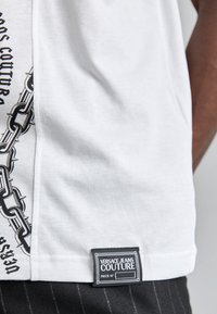 T-shirt en coton blanc avec un graphique de chaîne noire sur le côté et une étiquette portant "VERSACE JEANS COUTURE" à l'ourlet.