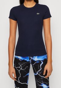 Žena v námořnickém pruhovaném tričku s krátkým rukávem a malým zeleným krokodýlem, doplněná černými kalhotami se vzorem modrých a bílých blesků.