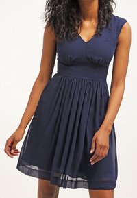 Navy blue dress with a fitted bodice, pleated skirt, and a v-neckline. Made from lightweight fabric with cap sleeves and a flared silhouette.