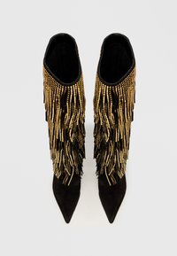 Crne nubuk čizme do gležnja s šiljastim prstima, ukrašene dugim zlatnim resicama za dodatnu teksturu i stil.