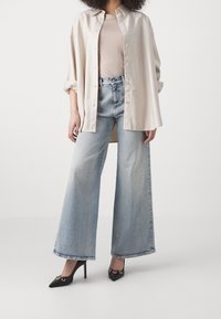 Camicia beige a righe con bottoni, maglietta rosa chiaro, jeans azzurri svasati e scarpe nere con punta a forma di stiletto dotate di dettagli dorati.