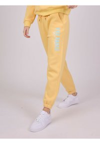 Gule sweatpants med elastiske manchetter og snor i taljen, der har blå tekst "PXP SQUAD" trykt på venstre side. Båret med hvide sneakers.