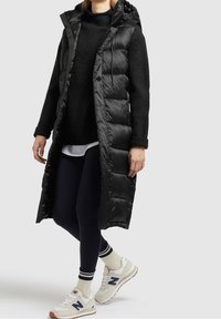 Czarna kurtka puchowa z kapturem, zatrzaskami i pikowanym wzorem; noszona na ciemnym swetrze, w towarzystwie granatowych legginsów i białych sneakersów.