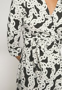 Svart omslagsklänning med ett vitt kedjemönster, med trekvartsärmar och ett knytmidjedetalj för en åtsittande form. Slät tygstruktur.