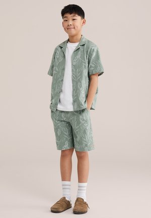 Garçon portant une chemise à manches courtes à motif de feuilles vertes et un short assorti, un t-shirt blanc, des chaussettes blanches et des chaussures marron sans lacets, debout les mains dans les poches.