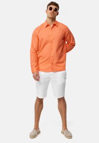 Orange langærmet skjorte i button-down stil, parret med hvide shorts og beige espadrilles. Model bærer runde solbriller.
