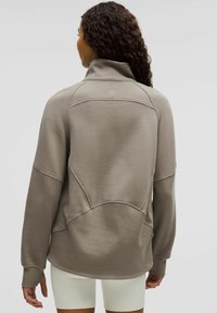 Lysebrun sweatshirt med høj krave, der har et tekstureret materiale, ribbede manchetter og et logo på ryggen. Designet inkluderer buede sømme.