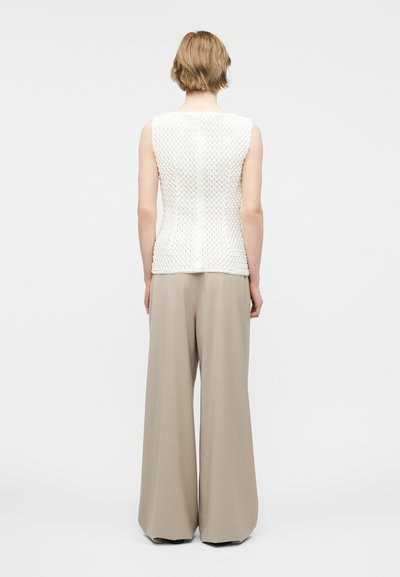 Άτομο με κοντά ξανθά μαλλιά, γυρισμένο με την πλάτη, φοράει λευκό αμάνικο τοπ με υφή και φαρδιά μπεζ παντελόνια σε απλό φόντο.