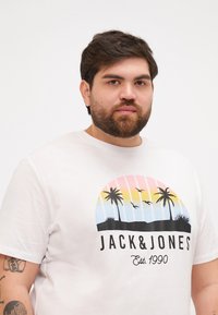 Hvid bomulds t-shirt med et farverigt grafisk motiv af palmer og fugle mod en solnedgangsbaggrund, med "JACK & JONES" tekst.