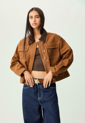 Veste en denim marron avec un col en velours côtelé, deux poches avant et des coutures blanches contrastantes, associée à un haut marron et un jean bleu.