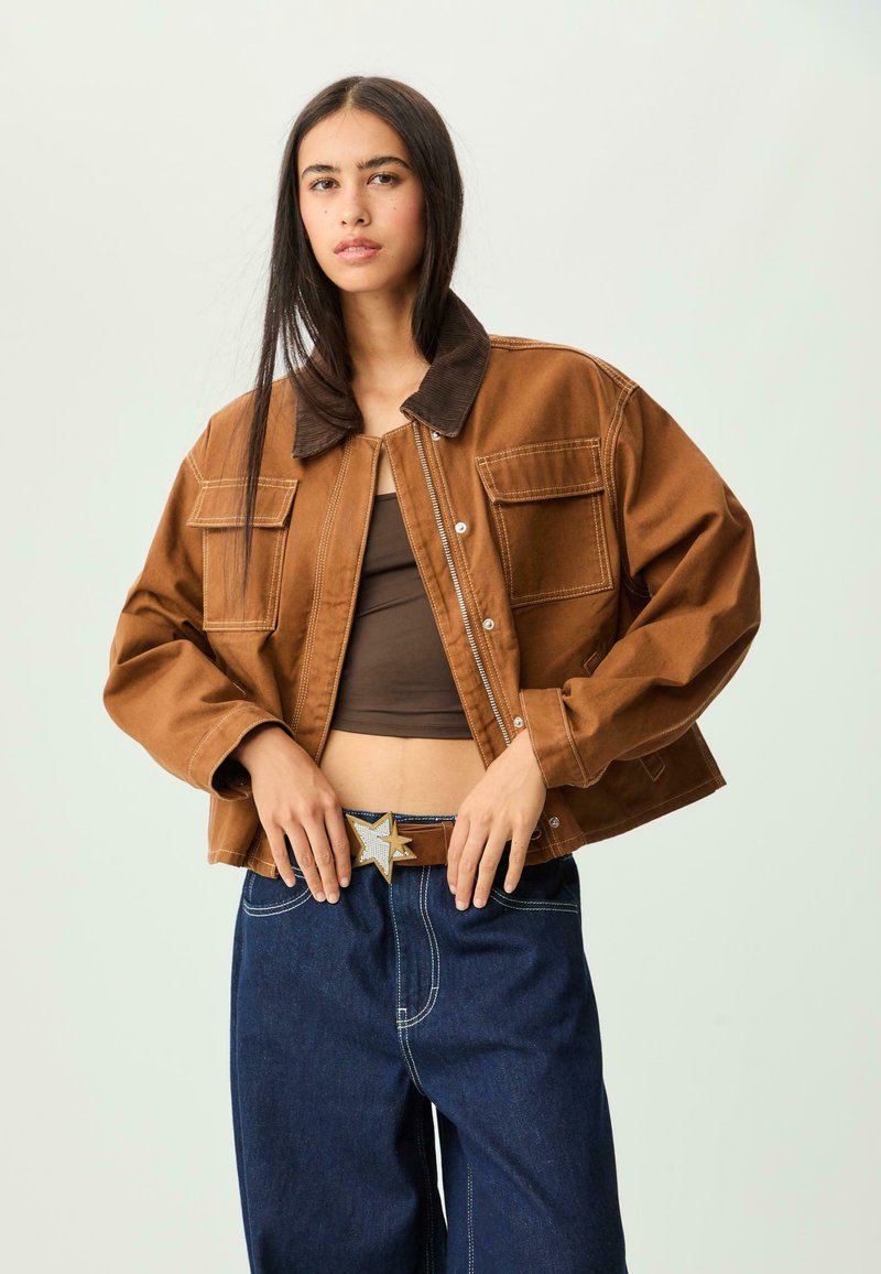 Brun denimjakke med et kordfløjelkrave, to frontlommer og kontrasterende hvid syning, parret med en brun top og blå jeans.