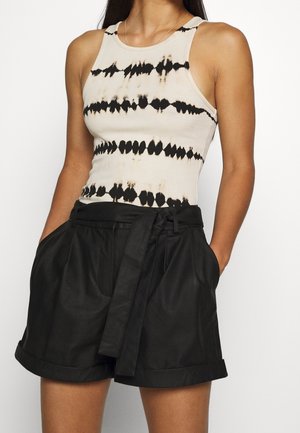 Vrouw draagt een mouwloze beige tie-dye tanktop en hooggetailleerde zwarte short met riem, handen in de zakken.