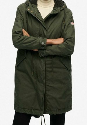 Personne portant une parka à capuche vert foncé avec poches avant et poignets boutonnés, les bras croisés, par-dessus un col roulé de couleur claire.