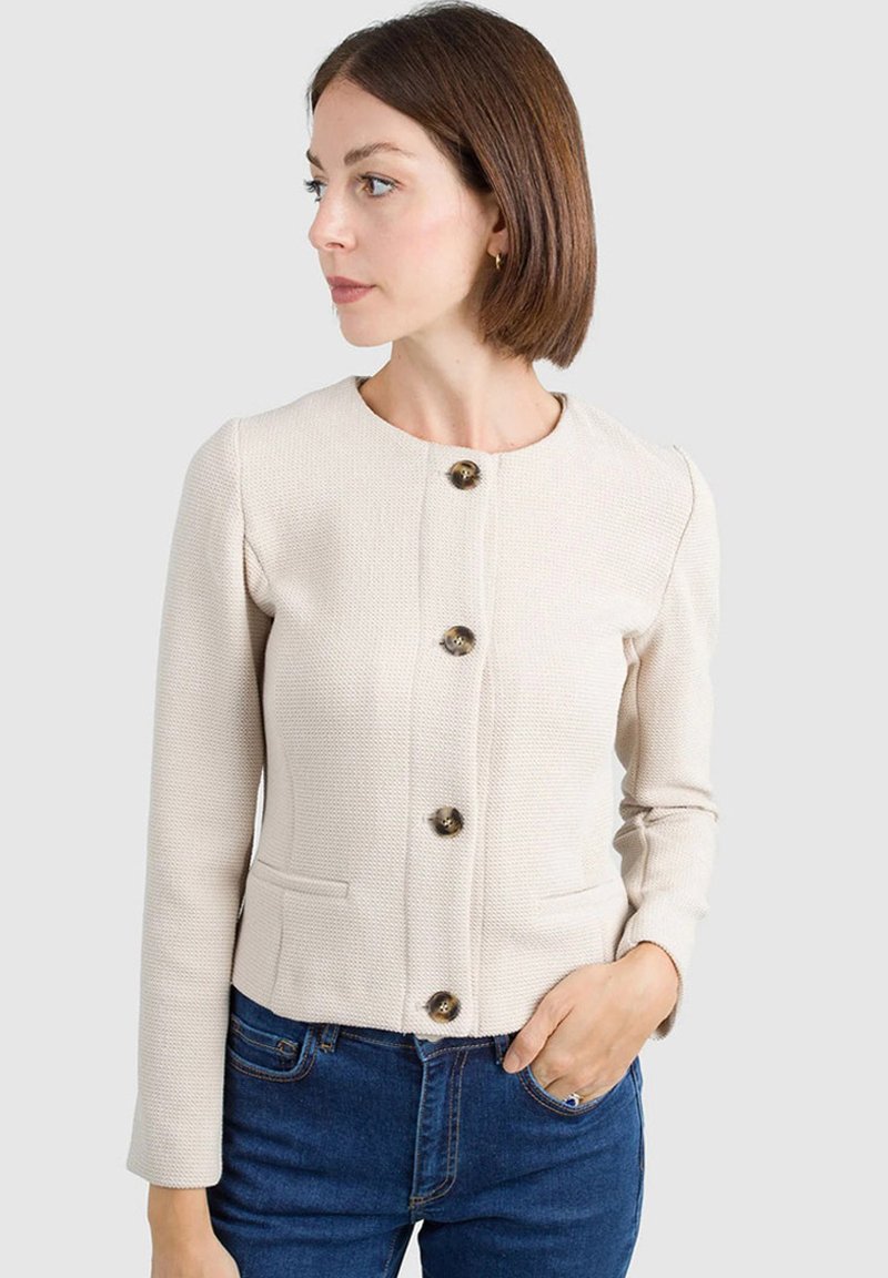 Giacca beige, texturizzata, con collo rotondo, maniche lunghe, chiusura con cinque bottoni e due tasche frontali. Indossata con jeans blu.