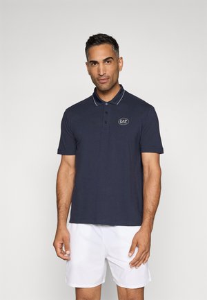 Marineblå polo med tre-knaps placket og kontrasterende hvid krave og manchetter. Har et broderet "EAZ" logo på brystet.