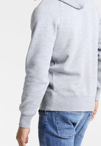 Mann in hellgrauer Kapuzenjacke und blauen Jeans, von hinten gezeigt mit leicht gebogenem linken Arm.