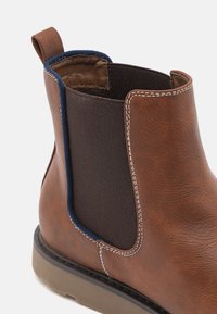 Brązowe skórzane kozaki za kostkę z elastycznym panelem bocznym, kontrastową niebieską podszewką i białym przeszyciem. Posiadają strukturalną powierzchnię i solidną podeszwę.