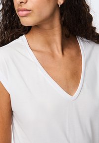 Kvinna med lockigt mörkt hår som bär en vit V-ringad topp och små hoopsörhängen, ser åt sidan.