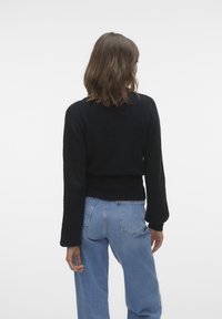 Svart stickad tröja med långa ärmar, ribbade muddar och en figursydd midja, kombinerad med ljusblå högmidjade jeans. Bakifrån.