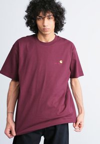Maroon oversized t-shirt made of cotton, featuring a small yellow logo on the chest and short sleeves. Smooth texture, casual fit.