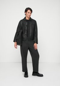 Casaco de cabedal preto com fecho de botão, usado sobre uma blusa preta. Calças largas cinza-escuras, sapatos pretos e uma mala tote preta.