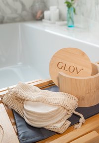 Pojemnik na bambus z logo "GLOV", bawełniane podkładki do twarzy i siateczkowe torby na drewnianej powierzchni, blisko kąpieli w stonowanych kolorach.