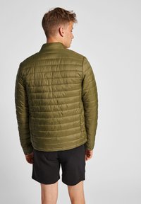 Grøn puffjakke med quiltet tekstur og ståkrave, iført over sorte shorts. Har synlige syninger og elastiske manchetter.