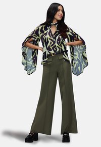 Blusa a vestibilità ampia, a fantasia in verde e viola con maniche a pipistrello, abbinata a pantaloni a gamba larga color oliva e scarpe con piattaforma nera chunky.