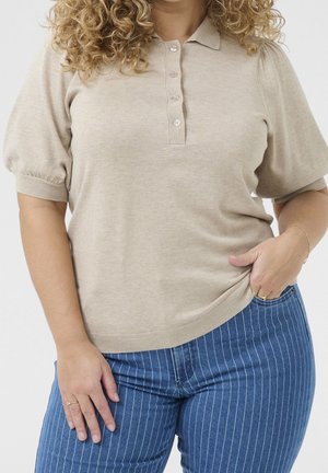 Kvinde med krøllet hår iført beige kortærmet strikket polo og blå bukser med nålestriber, hånd i lommen, mod ensfarvet lys baggrund.