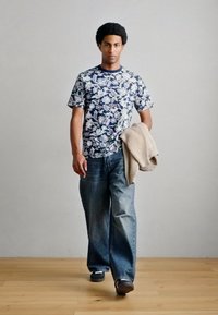 Μοντέλο που φοράει μια μπλε μπλουζά με φλοράλ σχέδιο και κοντά μανίκια, συνδυασμένη με χαλαρά ανοιχτό μπλε τζιν, κρατώντας ένα μπεζ πουλόβερ.